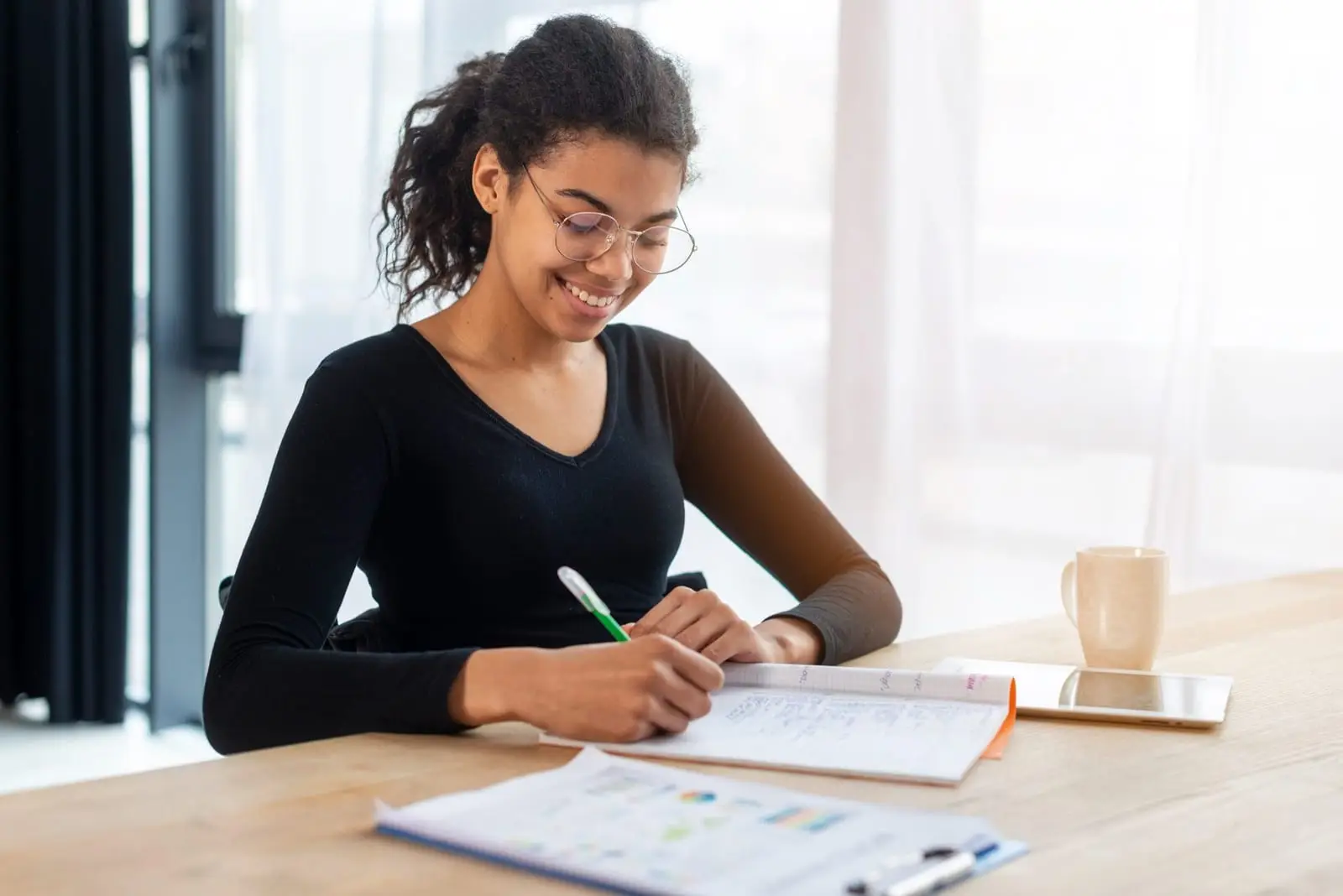 Estudante sorridente de óculos escrevendo em um caderno sobre uma mesa clara, com uma xícara ao lado
