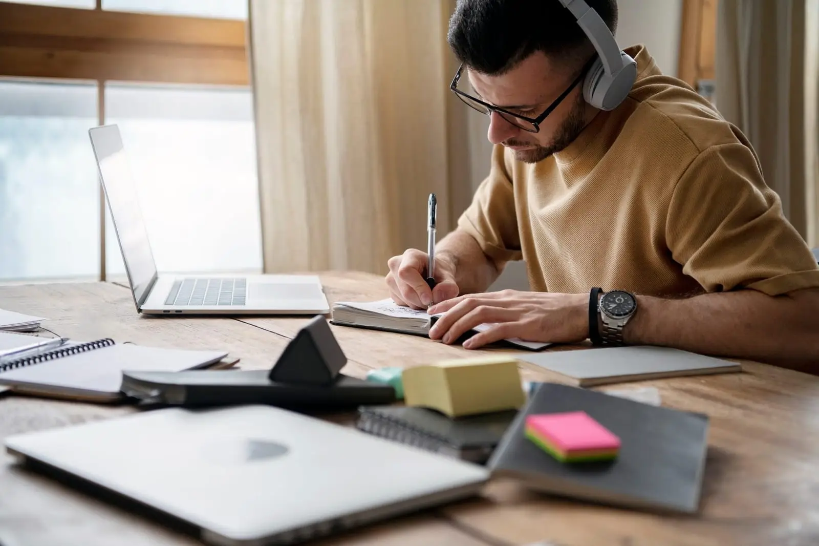 Estudante de fones de ouvido anotando em um caderno diante do notebook, com material de estudo espalhado pela mesa