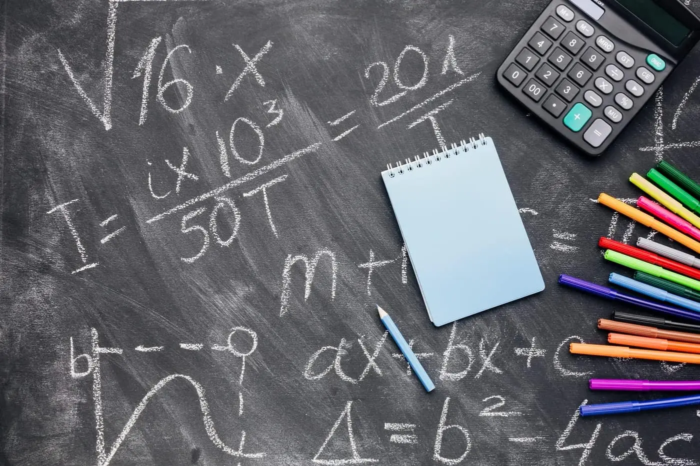 Quadro-negro coberto de equações escritas a giz, com calculadora, bloco de notas e canetas coloridas