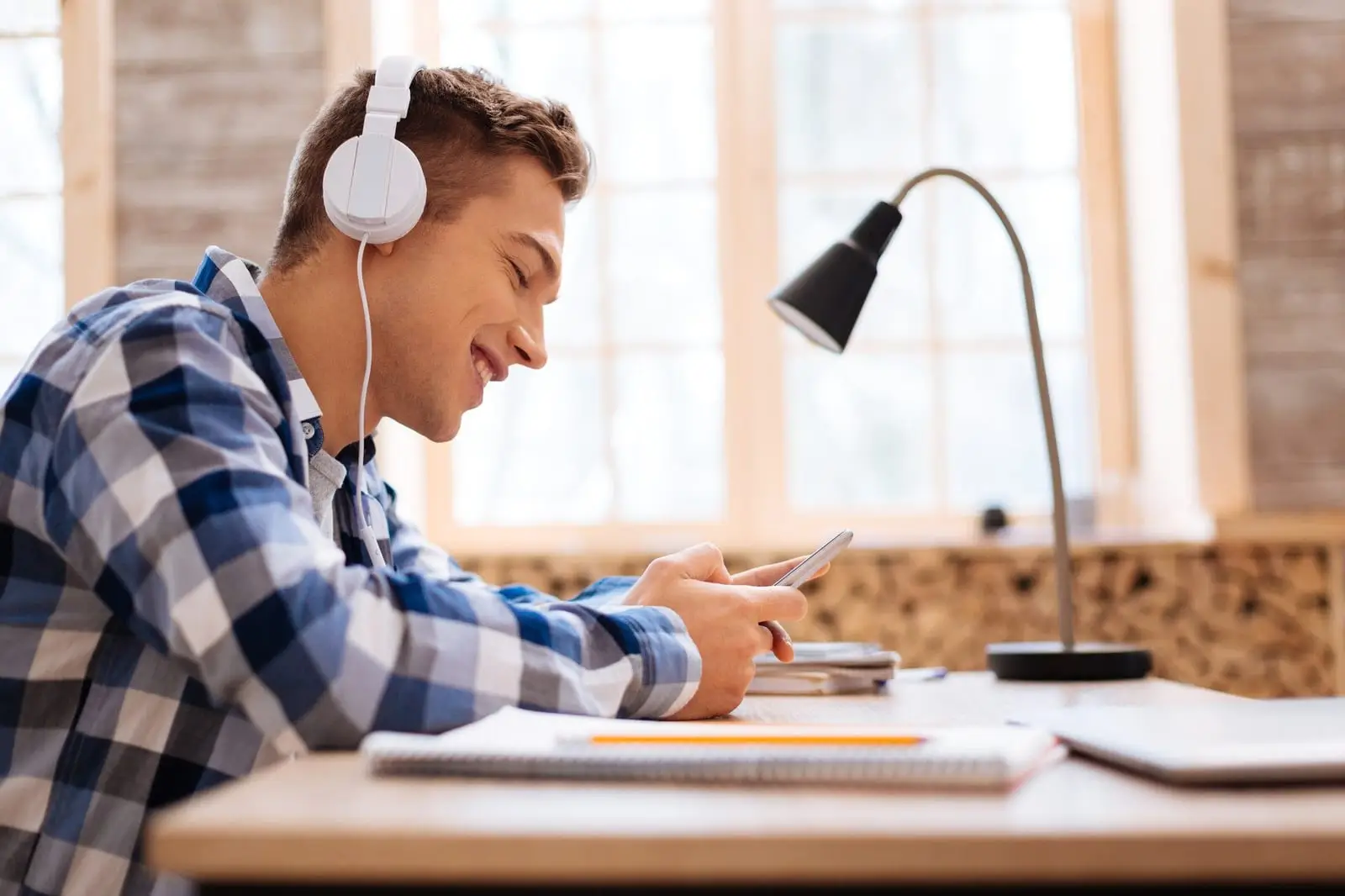 Estudante de fones de ouvido sorrindo enquanto escolhe uma música no celular, sentado à mesa de estudos